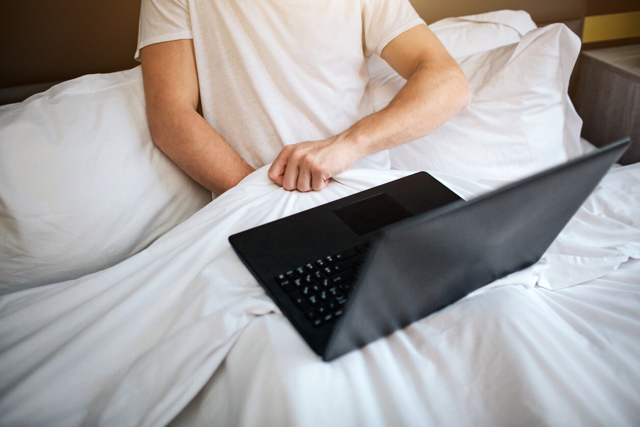 Young Man Sit in Bed Early Morning. He Hold Hand Under White Blanket and Masturbating. Laptop on His Legs.